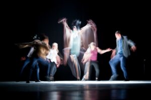 Five individuals, blurred by their motion, dancing around a sixth underneath a spotlight on stage. 